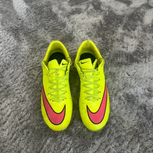 Gula Nike fotbollsskor med rosa swoosh - Säljer ett par riktigt snygga fotbollsskor från Nike i neon-gult med stora rosa swoosh-loggor på sidorna. Skorna har snörning och platta dobbar undertill för bra grepp på planen. Perfekta för dig som vill sticka ut på matchen!