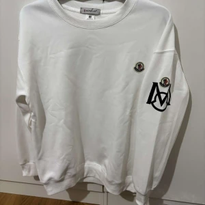 Vit Moncler sweatshirt med logga - Vit sweatshirt från Moncler med rund halsringning och ribbade muddar. Tröjan har Moncler-logga på bröstet och en stor svart Moncler-M på ena ärmen tillsammans med en extra logga. Skön och stilren modell i mjuk bomull.