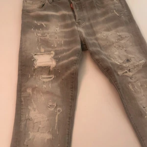Grå slitna skinny jeans från Dsquarred2 - Grå jeans från Dsquarred2 med skinny fit och coolt slitna detaljer, hål och blekta partier. Klassisk femficksmodell med dragkedja och knapp. Perfekta för dig som gillar en edgy streetstil.