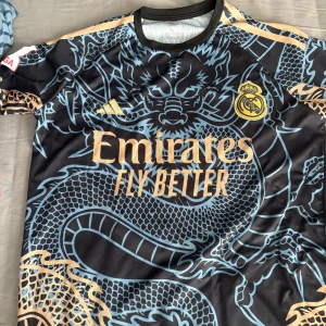 Real Madrid Bellingham fotbollströja - Snygg Real Madrid fotbollströja med Bellingham och nummer 5 på ryggen. Tröjan är svart med ljusblått och guldigt drakmönster, Real Madrid-emblem och Adidas-logga på bröstet. Tillverkad i lätt polyester med korta ärmar och rund hals.
