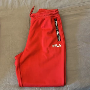 Röda mjukisbyxor från FILA - Snygga röda mjukisbyxor från FILA med logga på benet och dragkedjeficka med FILA-tryck. Byxorna har ribbad midja med snörning och är perfekta för en avslappnad stil. Tillverkade i mjukt material för maximal komfort.