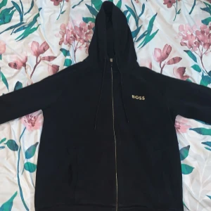 Svart hoodie från BOSS med dragkedja - Svart hoodie från BOSS i storlek L med guldfärgad BOSS-logga på bröstet. Tröjan har huva med snörning, hel dragkedja och två fickor framtill. Perfekt för en chill och stilren look.