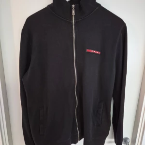 Svart ziptröja från Prada - Svart långärmad ziptröja från Prada med hög krage och diskret logga på bröstet. Tröjan har hel dragkedja framtill och två fickor. Tillverkad i mjuk bomull, perfekt för en clean och stilren look.