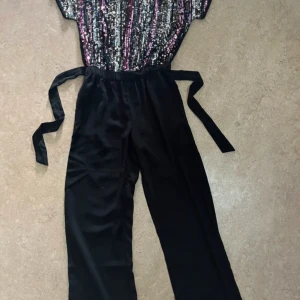 Svart byxdress med paljetter och knytband - Snygg svart byxdress med vida ben och knytband i midjan. Överdelen är täckt av glittriga paljetter i svart, silver och rosa för en extra festlig look. Byxorna är raka och tillverkade i ett mjukt tyg som ger en skön känsla.