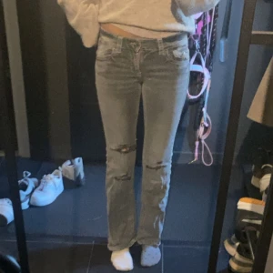 Grå bootcut jeans med slitningar - Säljer ett par gråa jeans från Gina  med bootcut passform. Jeansen har slitna detaljer på knäna och bakfickor med lock och knapp. Snygg tvätt och klassiska sömmar, perfekta för en avslappnad stil.