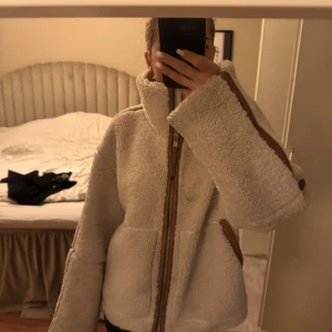 Beige teddyjacka med bruna detaljer - Supermysig beige teddyjacka med höga krage och bruna detaljer längs dragkedjan och ärmarna. Jackan har en oversized passform och är perfekt för kyliga dagar. Stängs med dragkedja framtill och har stora fickor på sidorna.
