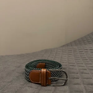 Grönvitt med brunt skinn  flätad bälte - Snyggt flätat bälte i mörkgrönt och vitt med bruna skinn-detaljer och silverspänne. Bältet har en cool, randig look och passar perfekt till jeans eller chinos. Den bruna läderänden ger en klassisk touch och det elastiska materialet gör det bekvämt att bära hela dagen.