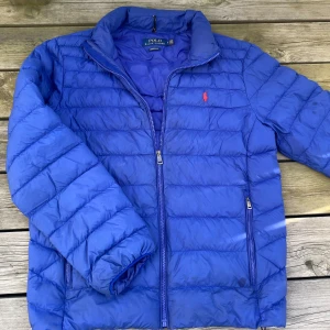 Blå dunjacka från Polo Ralph Lauren - Säljer en blå dunjacka från Polo Ralph Lauren med röd broderad logga på bröstet. Jackan har dragkedja framtill, två sidofickor. Perfekt för kyliga dagar och riktigt snygg till ett par jeans. Den har används fåtal gånger. Pris kan diskuteras!