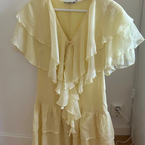 Ljusgul volangklänning från NA-KD - Supersöt ljusgul långklänning från NA-KD med volanger och v-ringning. Klänningen har knyt framtill och är tillverkad i ett lätt, luftigt material som känns perfekt för varma dagar. 