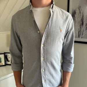Grå skjorta från Polo Ralph Lauren - Snygg grå skjorta från Polo Ralph Lauren i slim fit-modell. Skjortan har klassisk krage och tillverkad i mjuk bomull. Passar lika bra till fina tillfällen som till vardags. Killen på bilden är 178. 