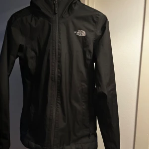 Svart North face jacka - Svart vindjacka från The North Face med huva och dragkedja framtill. Jackan har en stilren design med logga på bröstet och praktiska fickor med dragkedja. Perfekt för blåsiga dagar och enkel att matcha med allt.