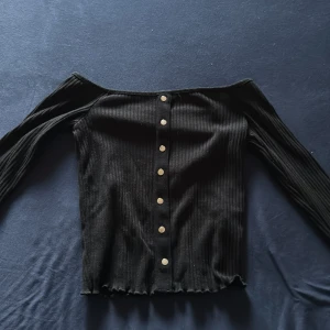 Svart ribbad offshoulder topp - Säljer en svart ribbad offshoulder topp med långa ärmar och dekorativa knappar framtill. Toppen har en croppad passform och vågig kant nertill.  Den är storlek Medium.