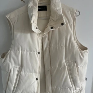 Vit quiltad dunväst från Selected - Snygg vit dunväst från Selected med hög krage och quiltad design. Västen har dragkedja och tryckknappar framtill samt två fickor på sidorna. Perfekt för lager-på-lager och ger en clean look till din outfit.