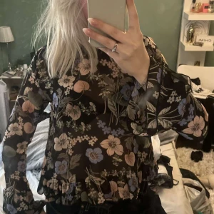 Blommig svart blus med volang - Säljer en transparent svart blus med blommigt mönster i lila, beige och grönt. Blusen har långa ärmar med volangdetaljer och en söt knytning i nacken. Perfekt för dig som gillar romantiska vibes och flowiga plagg.