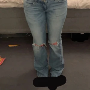 Ljusblå bootcut jeans med slitningar - Säljer ett par ljusblå jeans med bootcut passform och slitna detaljer vid knäna. Jeansen har klassiska fickor bak med lock och knapp, samt normal midja. 