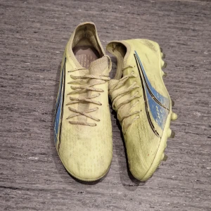 Puma Ultra gula elite AG - Säljer ett par gula Puma Ultra fotbollsskor med snörning och svarta detaljer. Skorna har en lätt ovandel i syntetmaterial och dobbar för spel på gräs. Snygg blå och svart design på sidan och texten ULTRA på hälen. Perfekta för dig som vill sticka ut på planen.