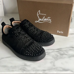 Svarta sneakers med nitar från Louboutin - Säljer ett par svarta sneakers från Christian Louboutin med coola svarta nitar på tån och klassisk röd sula. Skorna har snörning, rund tå och är tillverkade i mocka med läderfoder. Perfekt för dig som vill sticka ut med en edgy vibe. Två nitar på högra sko är borta och en ring på sidan av varje sko är borta och glapp i sidan på bägge sidor fram. Box och dustbag tillkommer!