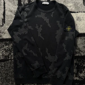 Stone island tröja - Svart sweatshirt från Stone Island med grått camoinspirerat mönster och klassisk patch på ärmen. Tröjan har rund hals, ribbade muddar och är tillverkad i mjuk bomull. Perfekt för dig som gillar streetwear och vill sticka ut med en unik design.