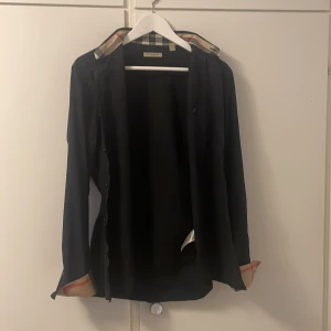 Svart skjorta från Burberry Brit - Stilren svart skjorta från Burberry Brit med klassiskt rutigt mönster på insidan av kragen och manschetterna. Skjortan har lång ärm, knappar framtill och en snygg, tidlös design. Perfekt för dig som gillar exklusiva detaljer och ikoniska mönster. nypris:4499