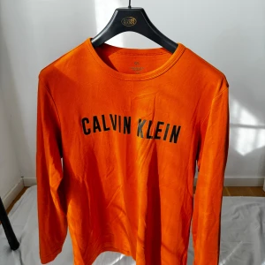 Orange långärmad tröja Calvin Klein - Snygg orange långärmad tröja från Calvin Klein med svart logotyptryck framtill. Tröjan har rund halsringning och är tillverkad i mjuk bomull, perfekt för dig som gillar en clean och sportig look. Storlek M