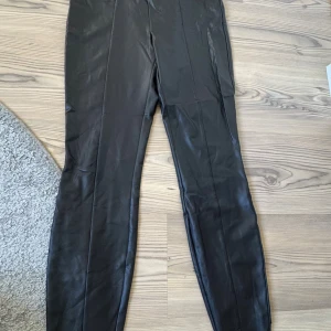 Svarta skinnbyxor från Vila - Snygga svarta skinnbyxor från Vila med hög midja och smal passform. Byxorna har en stilren look med raka sömmar framtill och är tillverkade i ett mjukt, syntetiskt skinnmaterial. Perfekta för dig som vill ha en edgy vibe i garderoben.