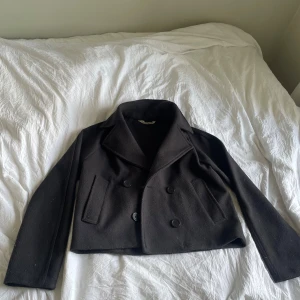Svart kappa från H&M - Säljer en svart kappa från H&M med dubbelknäppning och bred krage. Jackan har två snedställda fickor framtil.