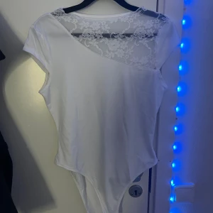Vit body med spetsdetalj och asymmetrisk axel - Säljer en vit body med korta ärmar och snygg asymmetrisk axel. Ovanpå axeln finns en transparent spets med blommönster som ger en extra touch. Perfekt att styla med jeans eller kjol :))