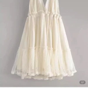 Ljus kortklänning med volang och knyt - Superfin ljus kortklänning med v-ringning, volangkant och knyt i ryggen. Klänningen är ärmlös och har ett skirt, prickigt tyg i flera lager som ger en romantisk känsla. Perfekt för sommarens varma dagar och har en luftig passform.