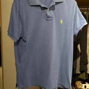Blå pikétröja från Ralph Lauren - Klassisk blå pikétröja från Ralph Lauren med korta ärmar och gul broderad logga på bröstet. Tröjan har krage och knäppning med två knappar. Perfekt för en clean och stilren look.