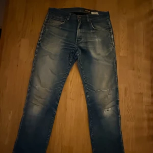 Blå slitna jeans från Wrangler ”Greenborg” - Säljer ett par blå jeans från Wrangler ”Greenborg” med snyggt slitna detaljer och ljus tvätt. Klassisk femficksmodell med läderpatch på bakfickan och raka ben. Perfekta för dig som gillar en avslappnad och trendig look.