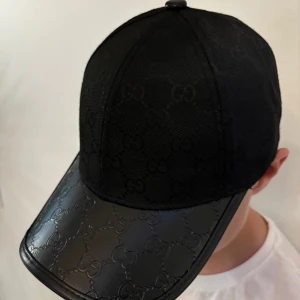Svart keps med GG-mönster - Säljer en svart keps med ikoniskt GG-mönster över hela kepsen och på skärmen. Skärmen är i blankt material med präglat mönster och kepsen har justerbar rem bak med gulddetalj. Perfekt för dig som vill ha en lyxig streetstyle-look.