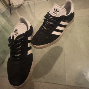 Adidas Gazelle svarta sneakers mocka - Klassiska Adidas Gazelle sneakers i svart mocka med vita detaljer och de ikoniska tre ränderna på sidorna. Skorna har vit plös och häl, svart snörning och gummisula. Perfekta för en stilren och sportig look. Skicka pris förslag 