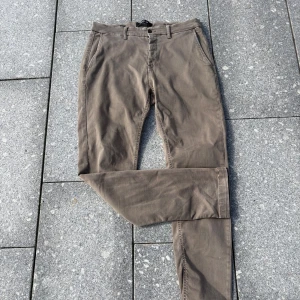 Beige Replay Hyperflex chinos - Snygga beige chinos från Replay Hyperflex med klassisk passform och raka ben. Byxorna har hällor för bälte, sidfickor och är tillverkade i ett stretchigt bomullsmaterial som ger extra komfort. Perfekta för en clean och stilren look.