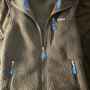 Patagonia väst - Mysig svart fleeceväst från Patagonia i storlek XS. Västen har en grov, teddy-liknande yta, tre fickor med dragkedja och blå detaljer vid dragkedjorna. Perfekt för lager-på-lager och outdoor-vibe. Klassisk Patagonia-logga på bröstet.