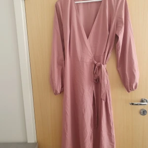 Ljusrosa omlottklänning med knyt - Säljer en lång, rosa omlottklänning med v-ringning och knyt i midjan. Den är aldrig använd och enbart testad en gång
