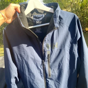 Marinblå Helly Hansen vindjacka M - Snygg marinblå vindjacka från Helly Hansen med Helly Tech Performance-tyg. Jackan har dragkedja framtill, hög krage och meshfoder på insidan. Perfekt för blåsiga dagar och har en clean look med diskret HH-logga på bröstet och ärmen. (NYPRIS 1759kr!!)