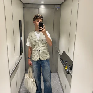 Beige utilityväst med dragkedja - Säljer en beige utilityväst med flera fickor och dragkedja framtill. Västen har en loose passform och är tillverkad i bomull. Perfekt för dig som gillar praktiska detaljer och streetstyle. Fickorna har både knappar och dragkedjor för extra funktion.