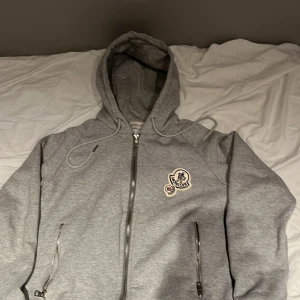 Grå kofta  med dragkedja - Snygg grå hoodie med dragkedja framtill, två fickor med dragkedja och broderad logga på bröstet. Tröjan har huva med snörning och är tillverkad i mjuk bomullsmix. Perfekt för en chill och stilren look.