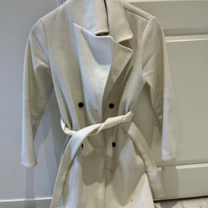 Beige trenchcoat med bälte - Snygg beige trenchcoat med klassisk krage och svarta knappar framtill. Jackan har ett matchande bälte i midjan för extra stilpoäng och långärmade ärmar. Materialet känns mjukt och har en ull-liknande struktur, perfekt för kyliga dagar.
