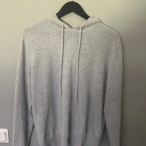 Ljusgrå Kashmir hoodie med huva - Mysig ljusgrå kashmir hoodie med huva och snörning. Tröjan har lång ärm och ribbade muddar vid ärmslut och nederkant. Perfekt för kyliga dagar när du vill vara både bekväm och snygg.