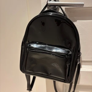 Svart glansig ryggsäck med dragkedja - Snygg svart ryggsäck i glansigt syntetmaterial med rundad form och praktisk framficka med dragkedja. Perfekt storlek för att få plats med det viktigaste och har justerbara axelremmar. Enkel och stilren design som passar till många outfits.