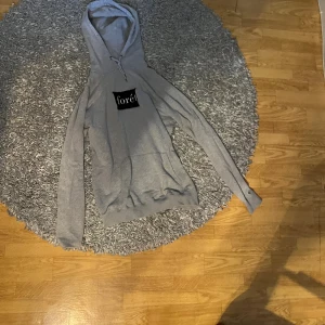 Grå hoodie från Forét med huva - Säljer en grå  hoodie från Forét i storlek L. Tröjan har en klassisk huva med snörning, stor magficka och svart Forét-logga på bröstet. Tillverkad i mjuk bomull, perfekt för chill dagar. Långärmad och loose fit för extra komfort.