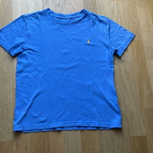 Blå t-shirt från Ralph Lauren - Blå t-shirt från Ralph Lauren med klassisk rund hals och korta ärmar. På bröstet finns den ikoniska broderade Polo-loggan i gult. T-shirten är gjord i mjuk bomull och har en clean, enkel look som funkar till jeans eller shorts.