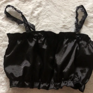 Svart satin  kort topp med volang - Säljer en svart kort topp i glansigt satinliknande material med smala axelband  med liten volangdetalj nedtill och upptill. Den är S men passar M, inte så stretchig tycker jag.