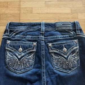Blå bootcut jeans från Miss Me - Snygga blå bootcut jeans från Miss Me med kontrastsömmar och detaljerade broderier på bakfickorna. Fickorna har coola vingar och stjärnor i silvertråd samt dekorativa knappar. Jeansen har klassisk femficksdesign och markerade sömmar för en extra edgy look. Köpta för ungefär 2200 kr. 