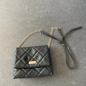 Svart quiltad axelväska med guldkedja - Snygg svart axelväska i quiltad design med guldfärgade kedjedetaljer och justerbar axelrem. Väskan har två stora fack och ett innerfack med dragkedja. Perfekt storlek för mobil, plånbok och andra essentials. Stilren och trendig look.