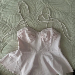 Ljusrosa topp med volang - Supersöt ljusrosa korsett-topp med tunna axelband och volang nertill. Kuporna är lätt rynkade för en snygg form och toppen har en feminin siluett. Perfekt att styla med jeans eller kjol för en trendig look.