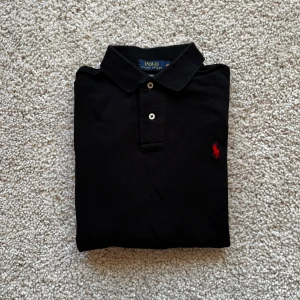 Svart tröja Ralph Lauren - Svart långärmad pikétröja från Polo Ralph Lauren i bomull. Klassisk krage, två knappar och röd broderad logga på bröstet. Custom fit-modell som sitter snyggt och stilrent. Perfekt för dig som gillar en clean och tidlös look. Skick 9/10🙏
