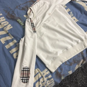 Vit Burberry sweatshirt med rutiga detaljer - Vit sweatshirt från Burberry med klassiska rutiga detaljer på axel och armbåge. Tröjan har rund hals, ribbade muddar och ett broderat Burberry-logo på bröstet. Tillverkad i mjuk bomull, perfekt för dig som gillar stilrena och ikoniska plagg.passar L och M 