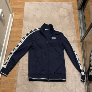 Blå Better Bodies vindjacka S - Snygg marinblå vindjacka från Better Bodies med vita detaljer och logga längs ärmarna. Jackan har dragkedja framtill, ribbade muddar och krage. Perfekt för dig som gillar sportig stil och vill ha något bekvämt och trendigt. Jackan har använts väldigt lite då den är förliten för mig. Den är i storlek Small för män, skriv vid funderingar